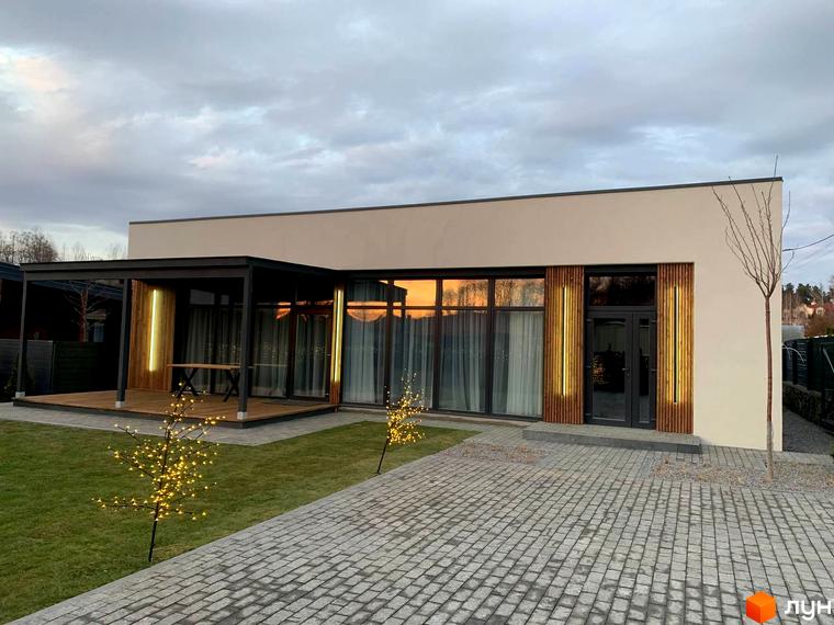 Котеджне містечко КМ Romankiv House. Будинок з панорамними вікнами та терасою, освітленою гірляндами, на тлі вечірнього неба. Прибудинкова територія з вимощеною доріжкою та декоративними деревами.