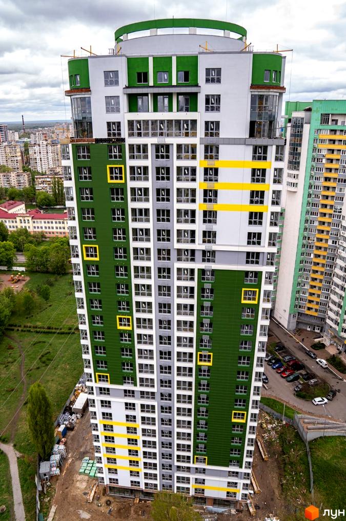 Високий житловий будинок Житлового комплексу Паркове місто з фасадом зеленого, білого та сірого кольорів, з жовтими акцентами. Навколо будинку видно інші споруди, зелені насадження та дорогу.