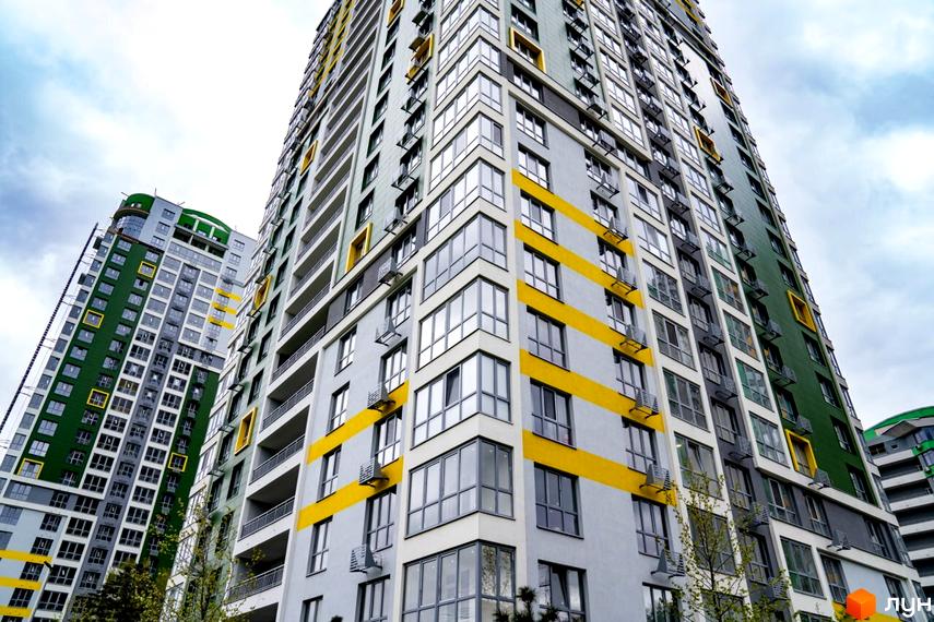 Фасад житлового комплексу Паркове місто з сірими стінами, жовтими смугами та зеленими вставками. На будинках багато вікон, балконів та видно дерева.