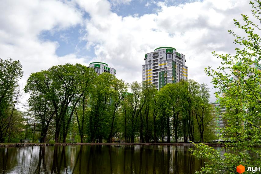 Житловий комплекс Паркове місто представлений висотними будинками сірого та зеленого кольорів з жовтими акцентами. Перед ними розташоване озеро, оточене густими зеленими деревами.