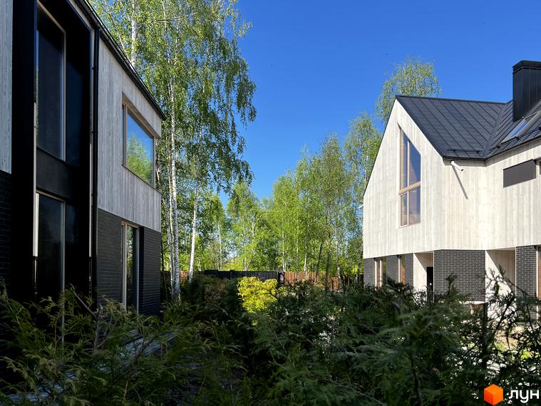 Котеджне містечко КМ Hidden House з дерев'яним фасадом та чорною цегляною основою на фоні зелені та блакитного неба. Фото новобудови.