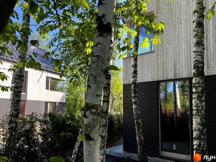 Зовнішній вигляд будинку КМ Hidden House з дерев'яною обшивкою та великими вікнами, оточений зеленню та деревами.