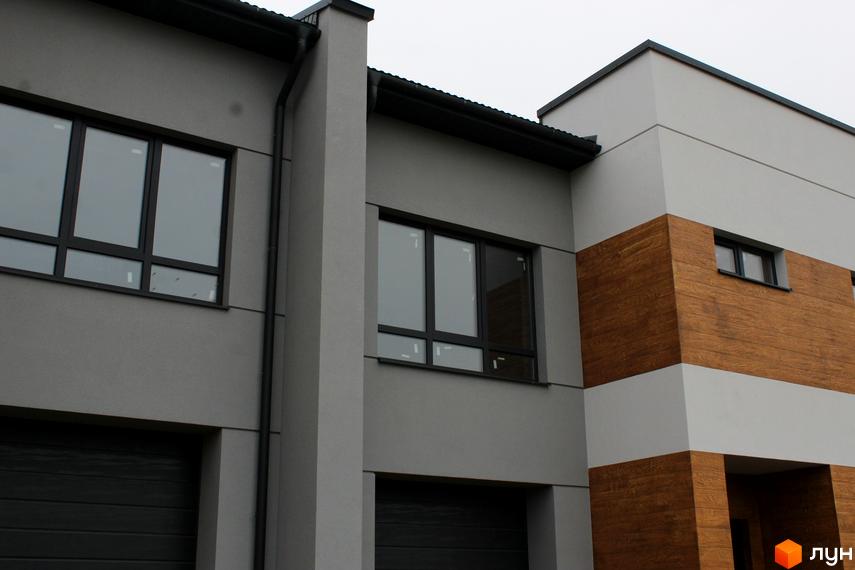 Зовнішній вигляд будинку в житловому комплексі Таунхаус „Пронятин“: сірі стіни, чорні вікна та гаражні ворота. Видно частину фасаду з дерев'яним оздобленням.