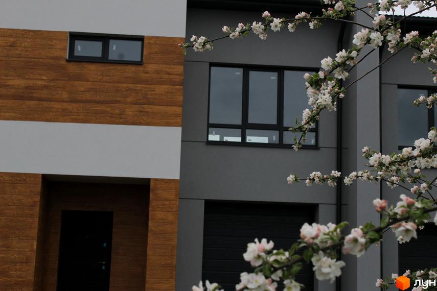 Фасад будинку з дерев'яним оздобленням та великими вікнами. На передньому плані квітучі гілки дерев.