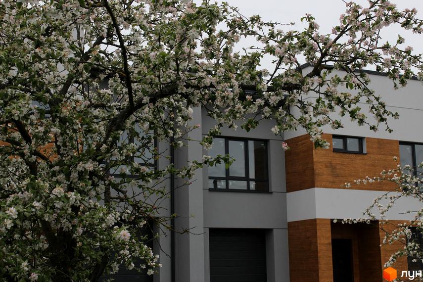 Таунхаус „Пронятин“ з сірим фасадом та коричневими вставками, частково прихований квітучим деревом.