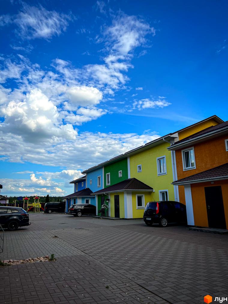Таунхауси Ivankiv-townhouse: яскраві фасади будинків у блакитному небі з хмарами. Прибудинкова територія з бруківкою та автомобілями.