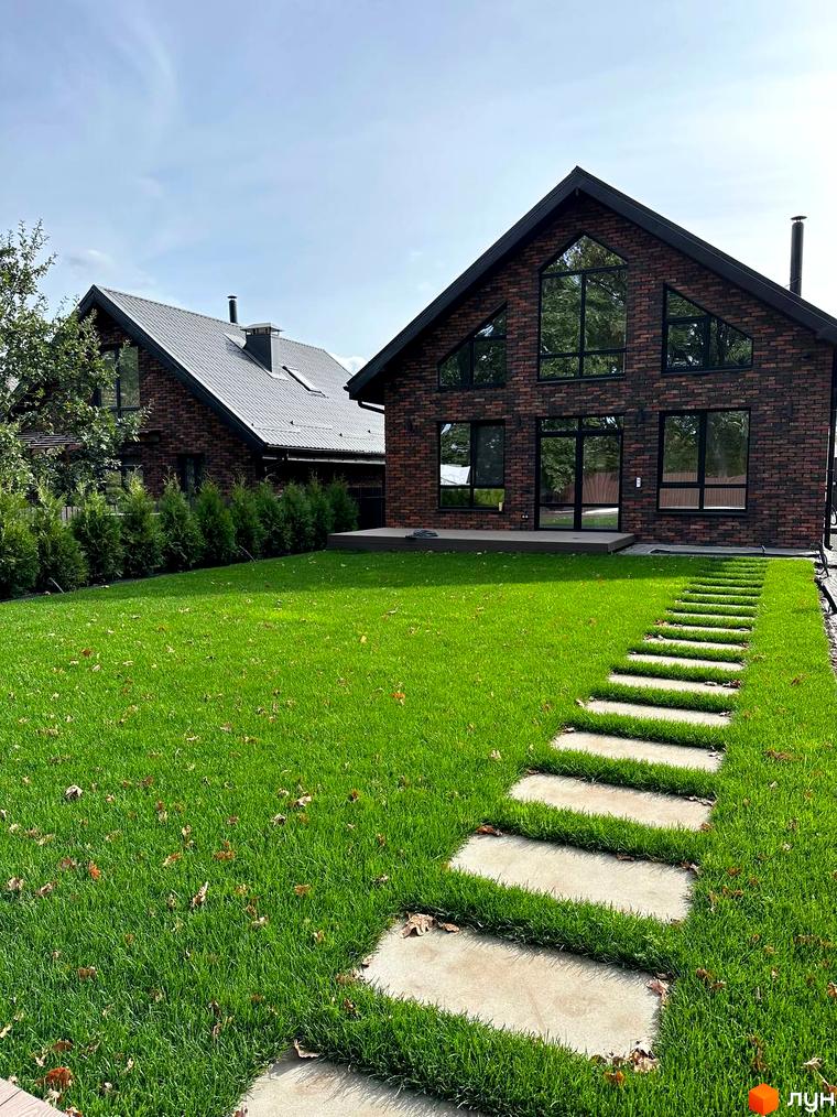 Цегляний будинок з великими вікнами та чорним дахом в Містечко котеджів Південна Баварія. Доглянутий газон з доріжкою з плитки, що веде до входу.