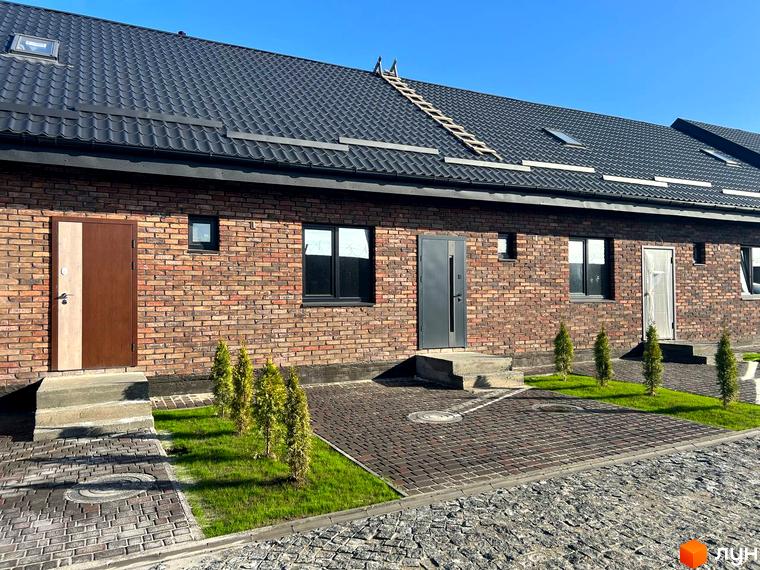 Цегляний будинок з темною покрівлею та дверима. На прибудинковій території бруківка та зелені насадження. КМ Південна Баварія - вигідна інвестиція в нерухомість.