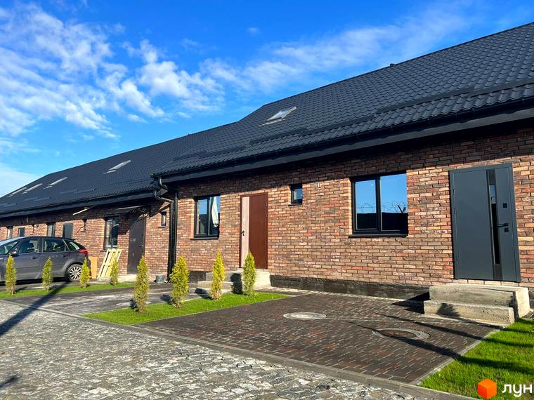 Цегляний будинок з темним дахом в Котеджному містечку Південна Баварія. Бруківка на подвір'ї та зелені насадження додають затишку.