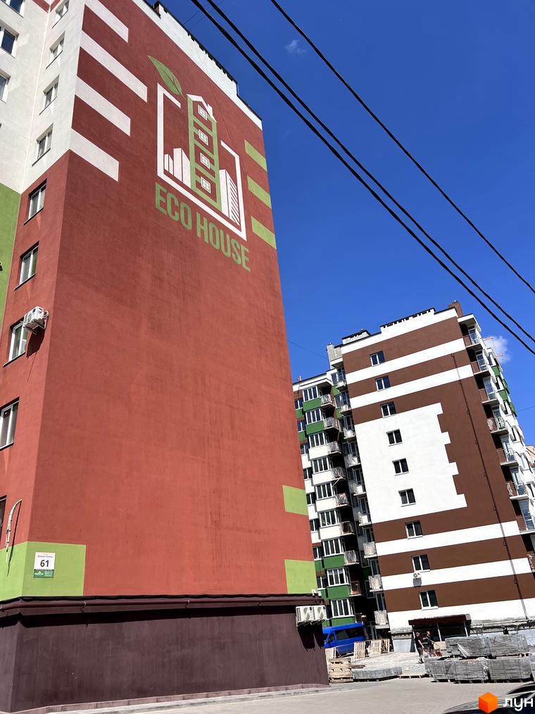 Житловий комплекс Eco House з червоним фасадом та біло-зеленими смугами поруч з коричнево-білою новобудовою. Навколо будівельні матеріали та синє небо.