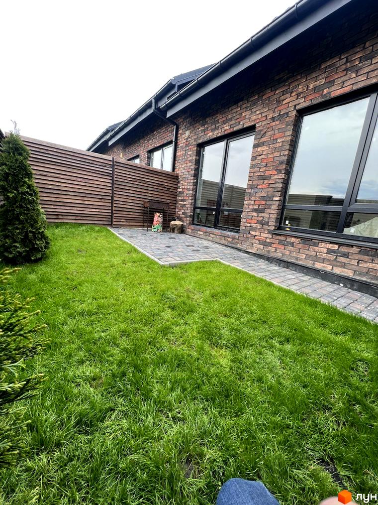 Котеджне містечко Одноповерховий Київ - Гатне. Фасад будинку з червоної цегли, великі вікна, зелений газон та дерев'яна огорожа.