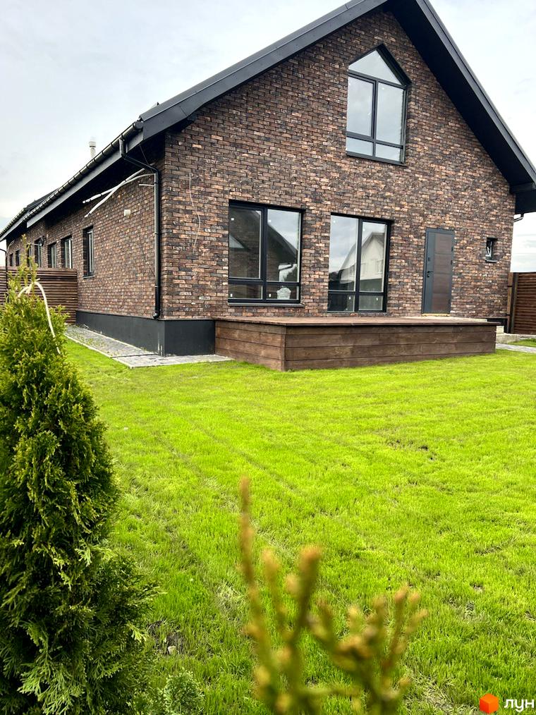 Котеджне містечко КМ Одноповерховий Київ - Гатне. Цегляний будинок з великими вікнами та дерев'яною терасою на тлі зеленого газону.