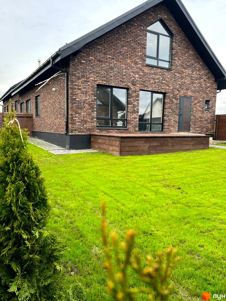 Котеджне містечко Квартал Баварія: двоповерховий будинок з цегляним фасадом, великими вікнами та дерев'яною терасою на тлі зеленого газону.