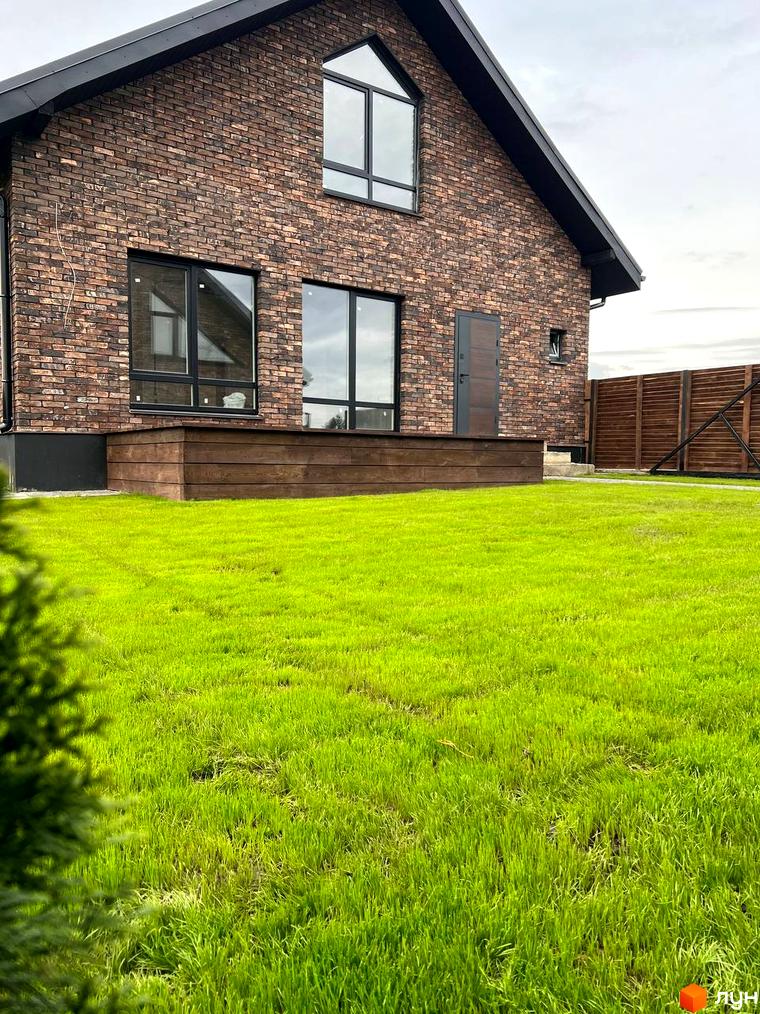 Котеджне містечко КМ Квартал Баварія: цегляний будинок з великими вікнами та доглянутою зеленою галявиною перед ним. На території розташована дерев'яна тераса та паркан.