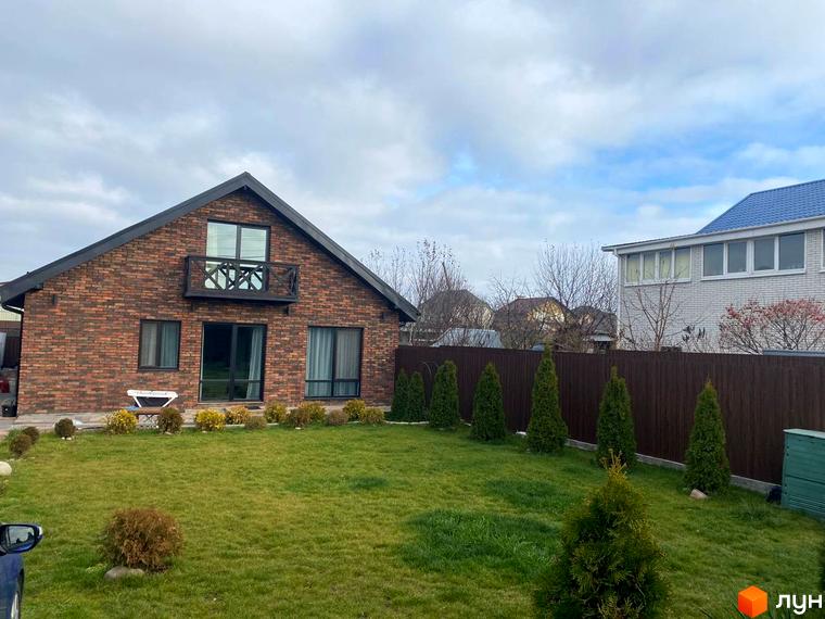 Одноповерховий будинок з червоної цегли з балконом та зеленим газоном навколо. Поруч розташований інший будинок та дерев'яний паркан.