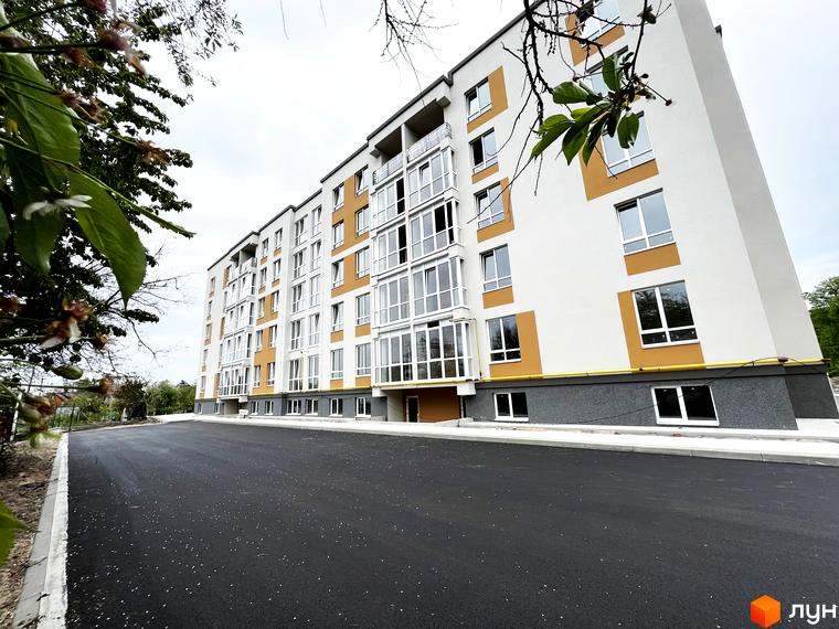 Зовнішній вигляд житлового комплексу Сімейний з фасадом білого та жовтого кольорів та великою кількістю вікон. Прибудинкова територія з асфальтованим покриттям.