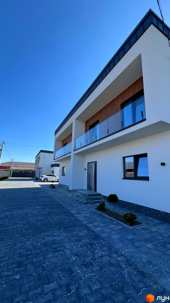 Двоповерховий будинок з білими стінами, скляними балконами та темною покрівлею. Бруківка на подвір'ї та зелені насадження.