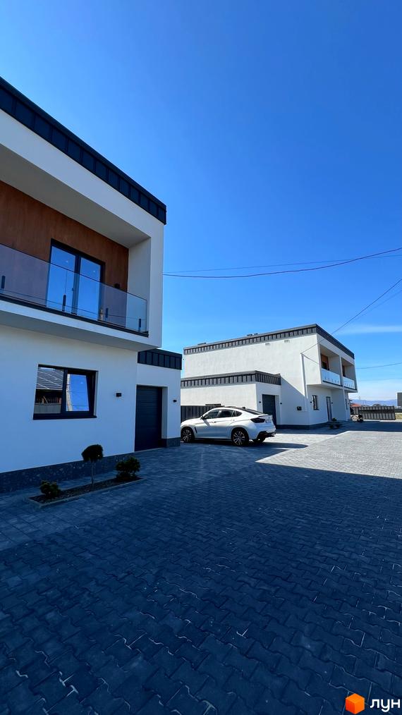 Двоповерхові будинки з білими фасадами та скляними балконами. На подвір'ї бруківка та припаркований автомобіль. Twins House - ідеальний вибір для комфортного життя.