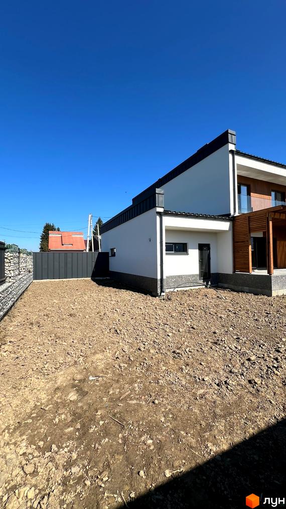 Білосніжний будинок з темним дахом та дерев'яними елементами фасаду. Поряд з будинком земляна ділянка та кам'яний паркан.