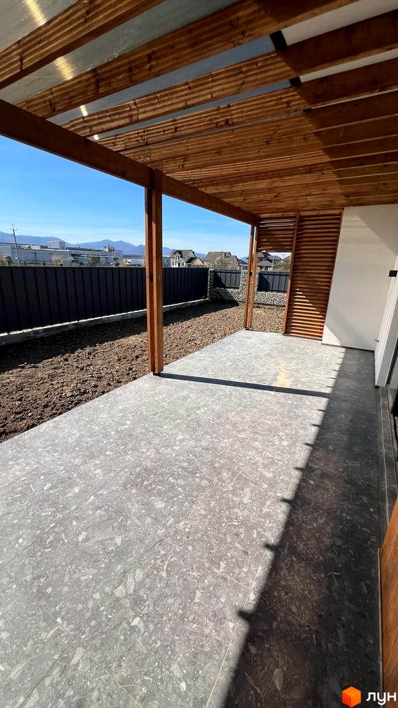 Простора тераса з дерев'яною стелею та підлогою з плитки. На задньому плані огорожа та зелена зона, що примикає до будинку.