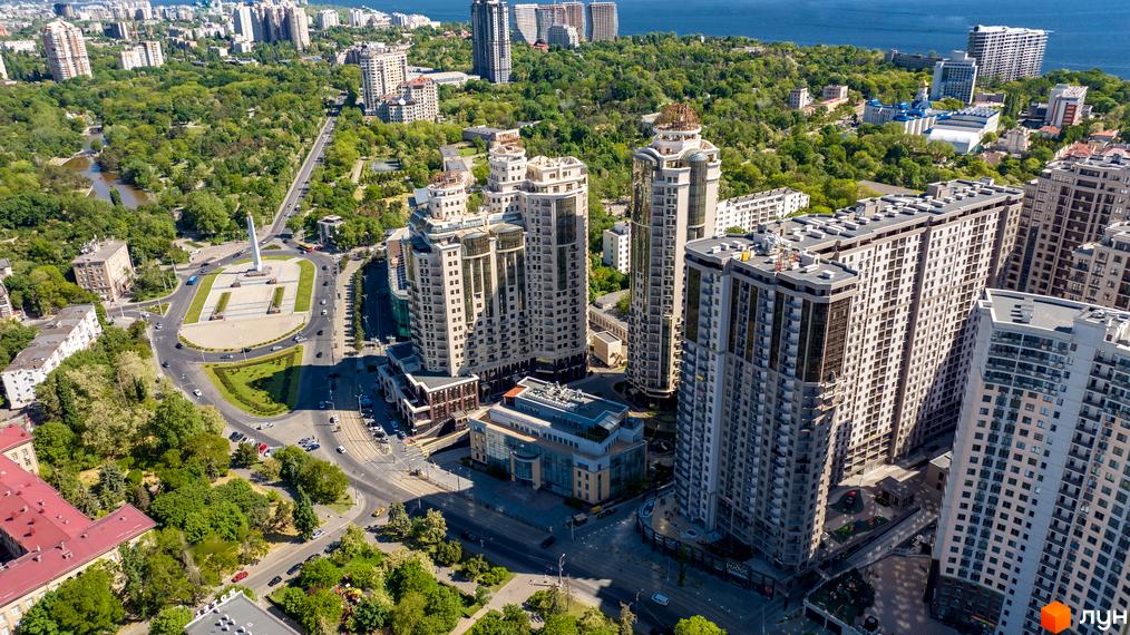 Аерофото житлового комплексу Родос: світлі багатоповерхові будинки з балконами. Поруч зелені зони, дороги, міська забудова та вид на море.