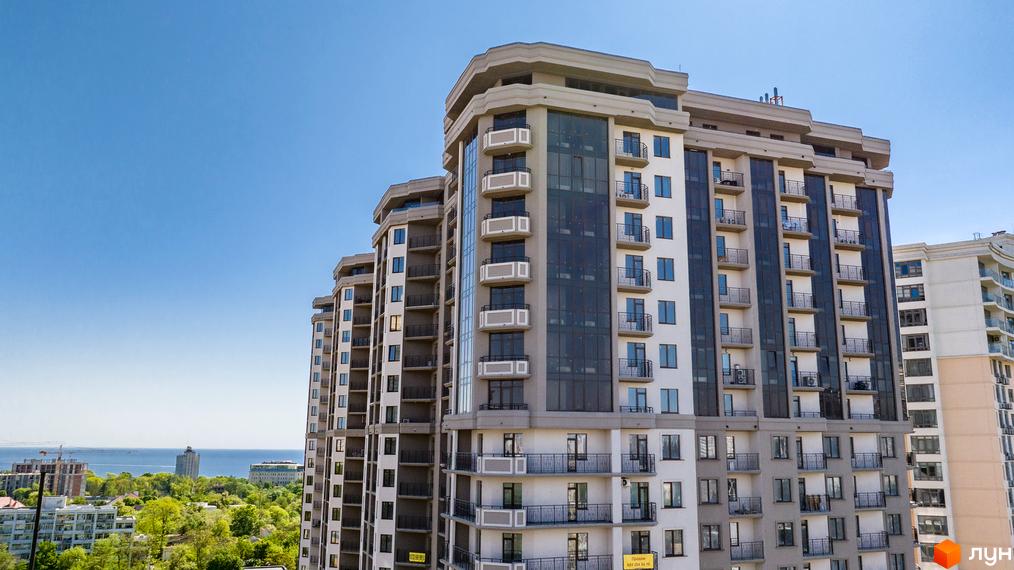 Багатоповерховий житловий комплекс Родос з світлим фасадом та темними скляними балконами. Навколо зелені дерева та вид на море.