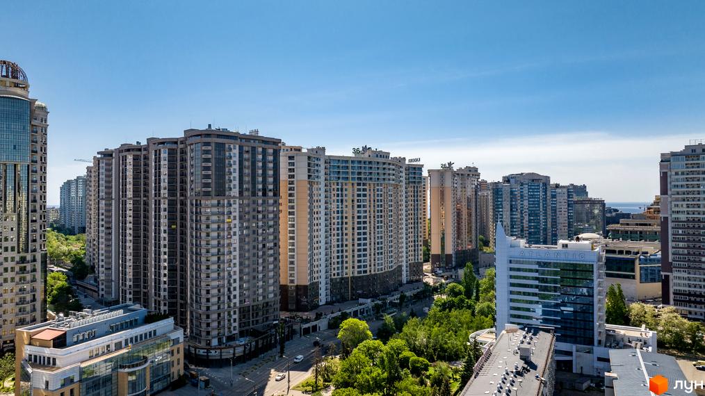 Висотні будівлі Житлового комплексу Родос з різнокольоровими фасадами та балконами. Поруч зелені насадження, дорога з автомобілями та вид на море.