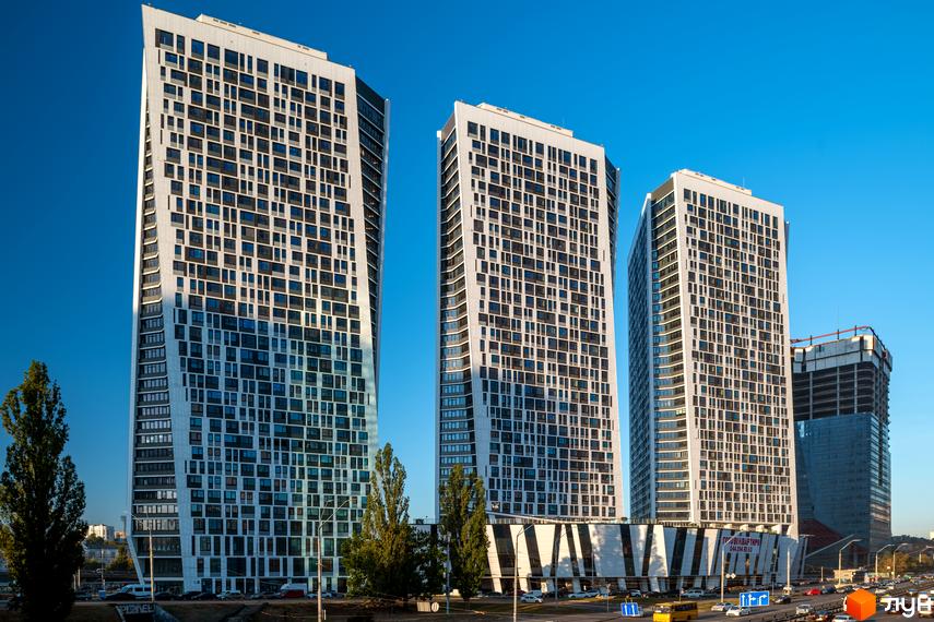 Житловий комплекс Manhattan City - три високі білі вежі з численними вікнами та вигнутими фасадами на тлі блакитного неба. На передньому плані дорога з автомобілями та зелені насадження.