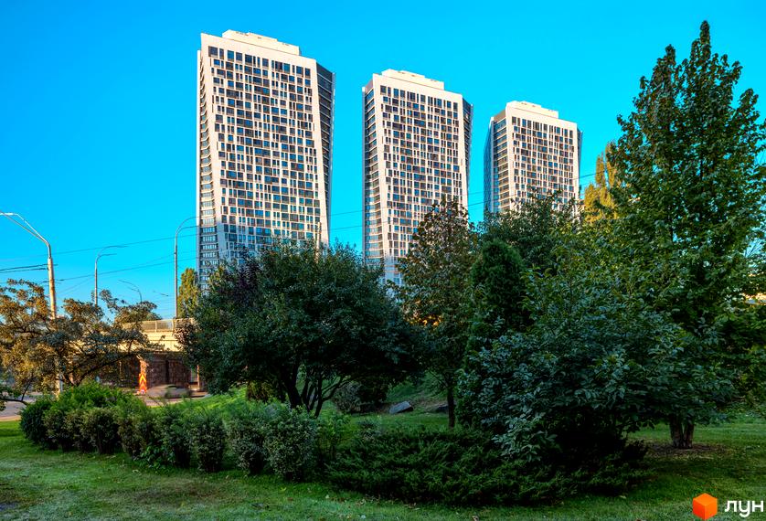 Житловий комплекс Manhattan City в Києві. Три висотні білі будівлі з численними вікнами на фоні блакитного неба. Перед будинками зелена зона з кущами та деревами.