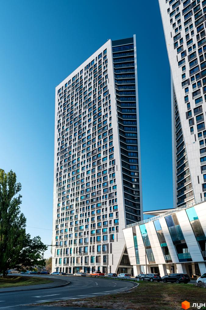 Житловий комплекс Manhattan city – високі білі вежі з синіми вікнами на тлі блакитного неба. Поруч дорога з автомобілями та зелені дерева.
