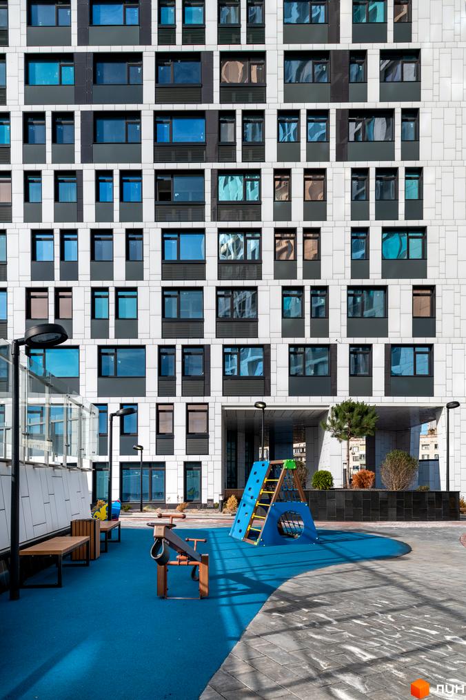 Фасад житлового комплексу Manhattan city з великою кількістю вікон та балконів. Поруч дитячий майданчик з гіркою та гойдалкою на синьому покритті.