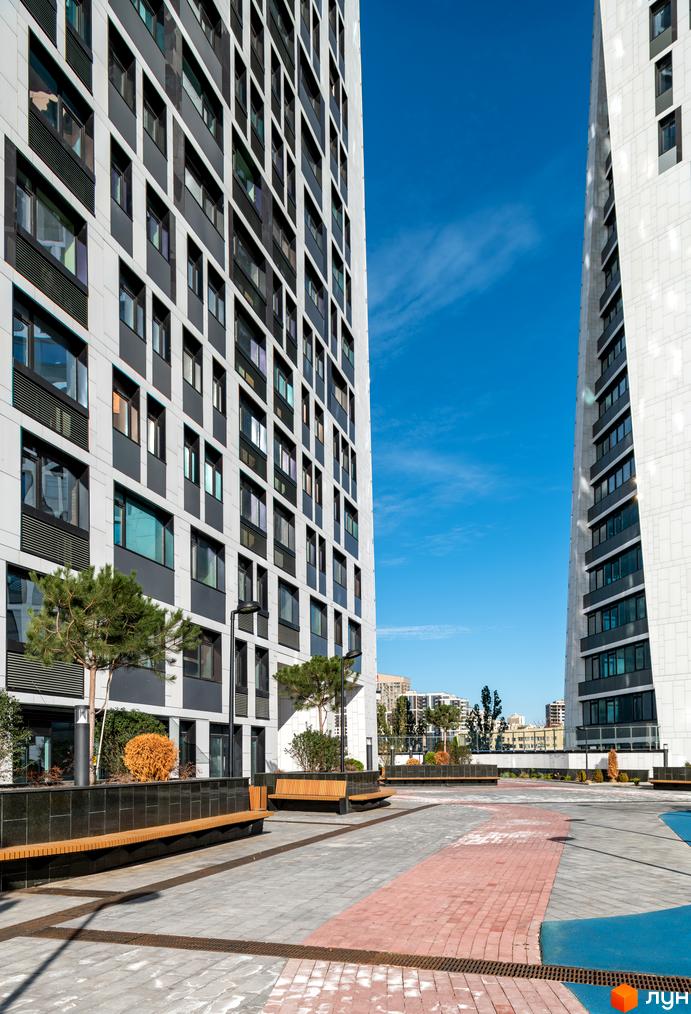 Житловий комплекс Manhattan city з біло-сірими фасадами та великою кількістю вікон. На прибудинковій території розташовані лавки та зелені насадження.