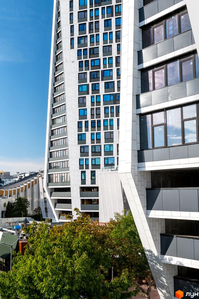 Новобудова ЖК Manhattan city з біло-сірим фасадом та синіми вікнами на тлі блакитного неба. На передньому плані зелені дерева.