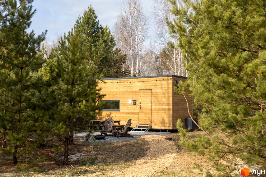 Дерев'яний будинок з панорамним вікном, оточений соснами. Біля будинку розташовані два крісла та мангал.