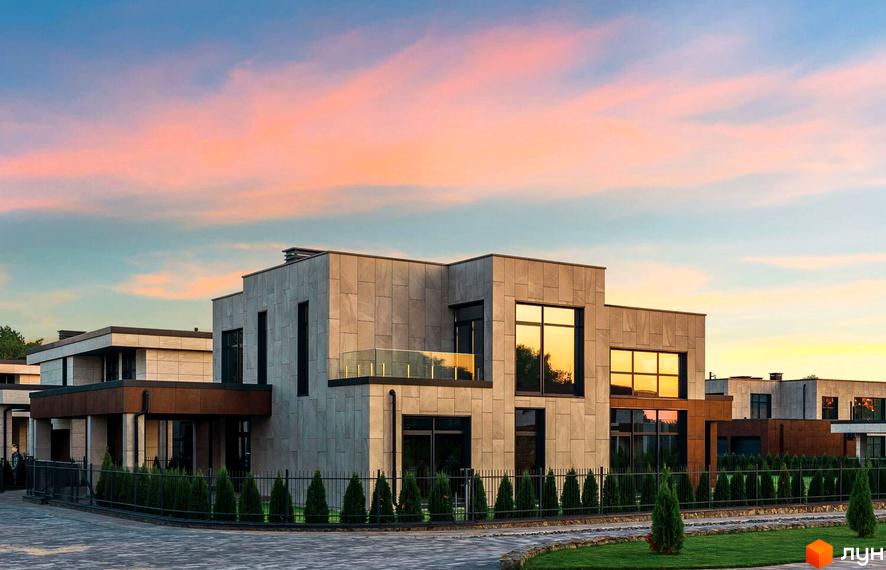 Котеджне містечко КМ DAMBA RIVER PARK з фасадами з натурального каменю та великими вікнами на тлі вечірнього неба. Озеленена прибудинкова територія з парканом та доглянутими туями.