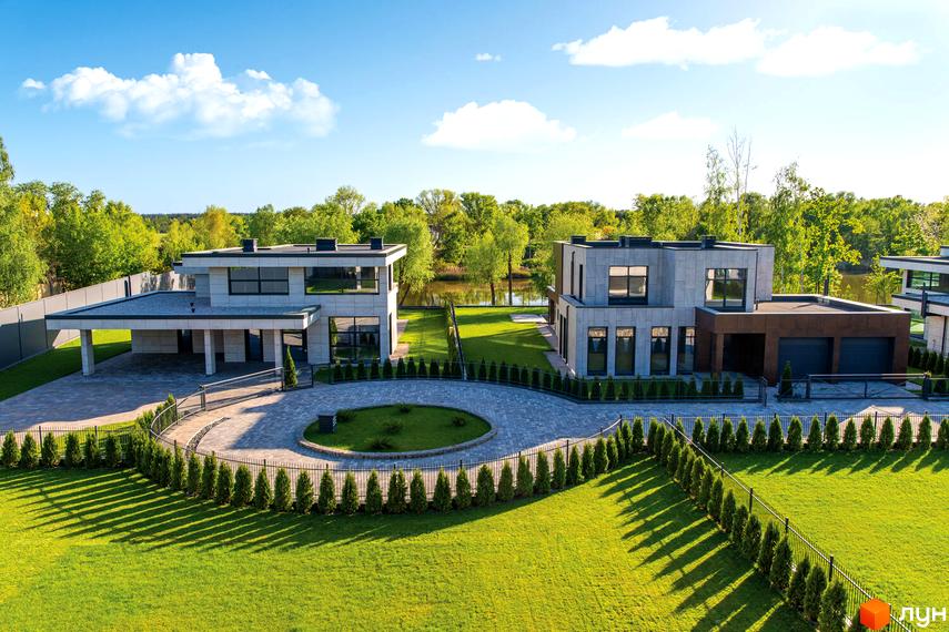 Котеджне містечко DAMBA RIVER PARK. Два двоповерхових будинки з сірою облицовкою та великими вікнами розташовані на доглянутій ділянці з газоном та огорожею.