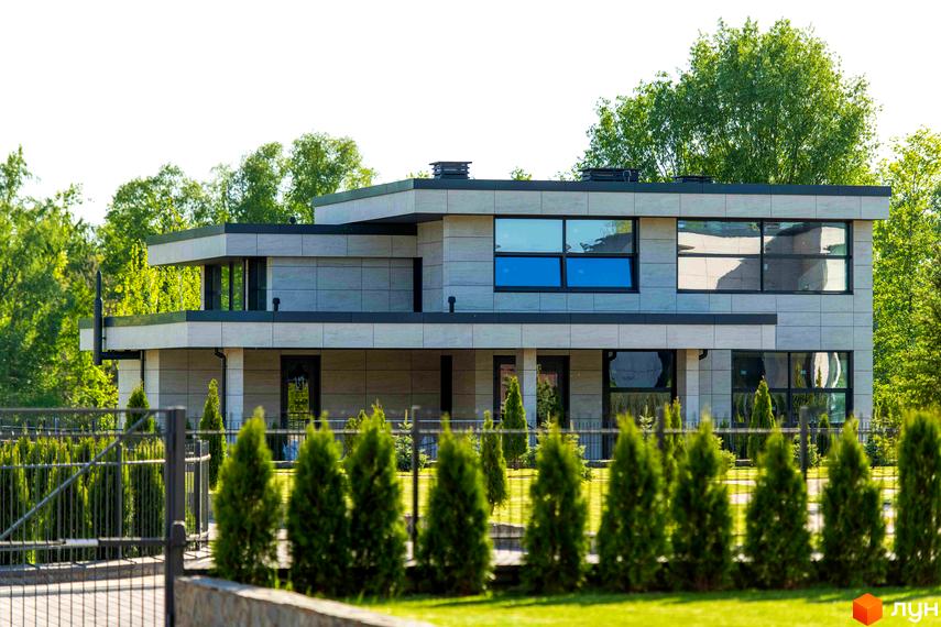 Котеджне містечко КМ DAMBA RIVER PARK. Фасад будинку оздоблений плиткою, великі вікна, навколо зелені туї та огороджена територія.