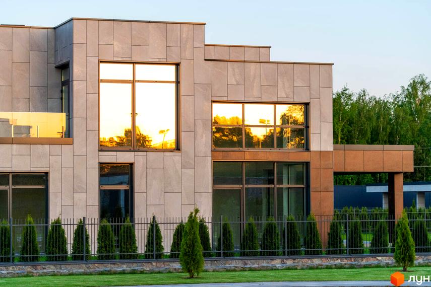 Котеджне містечко КМ DAMBA RIVER PARK. Фасад будинку з великими вікнами, що відбивають сонячне світло, та доглянута прибудинкова територія з газоном і огорожею.