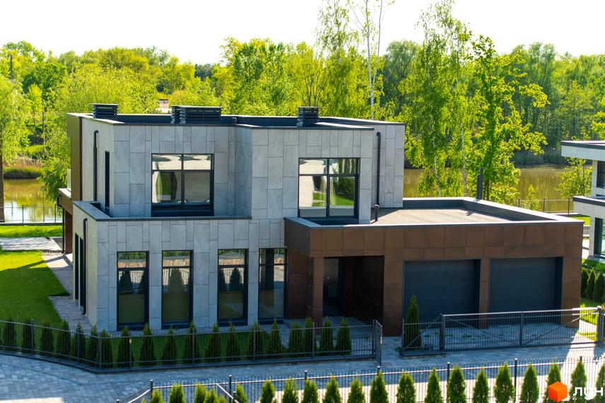 Котеджне містечко КМ DAMBA RIVER PARK. Сучасний двоповерховий будинок з сірим фасадом, великими вікнами та коричневим гаражем на два автомобілі.