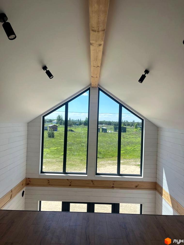 Котеджне містечко Залісся Village з двома великими вікнами, що виходять на зелене поле та невеликі будівлі. Інтер'єр з дерев'яними балками та білими стінами.