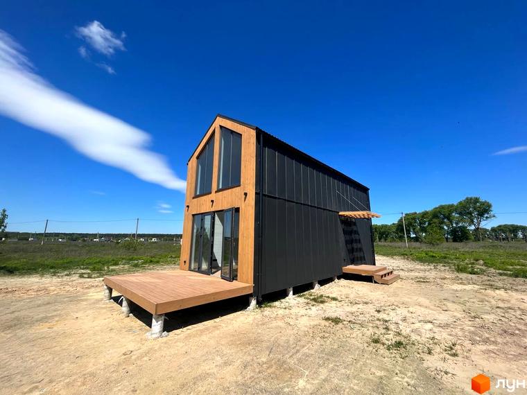 Котеджне містечко Залісся Village: двоповерховий будинок з панорамними вікнами та дерев'яною терасою на тлі блакитного неба. Навколо будинку – зелена трава та піщана ділянка.