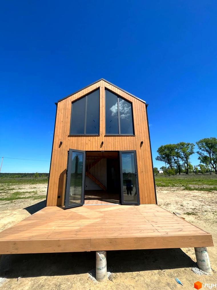 Котеджне містечко Залісся Village: двоповерховий дерев'яний будинок з великими вікнами та терасою на тлі блакитного неба та зелених дерев.