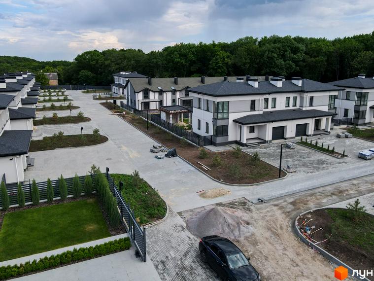 Зображення фасаду будинку в Котеджному містечку Ecopark з білими стінами та темним дахом. На території видно дорогу, зелені насадження та припаркований автомобіль.