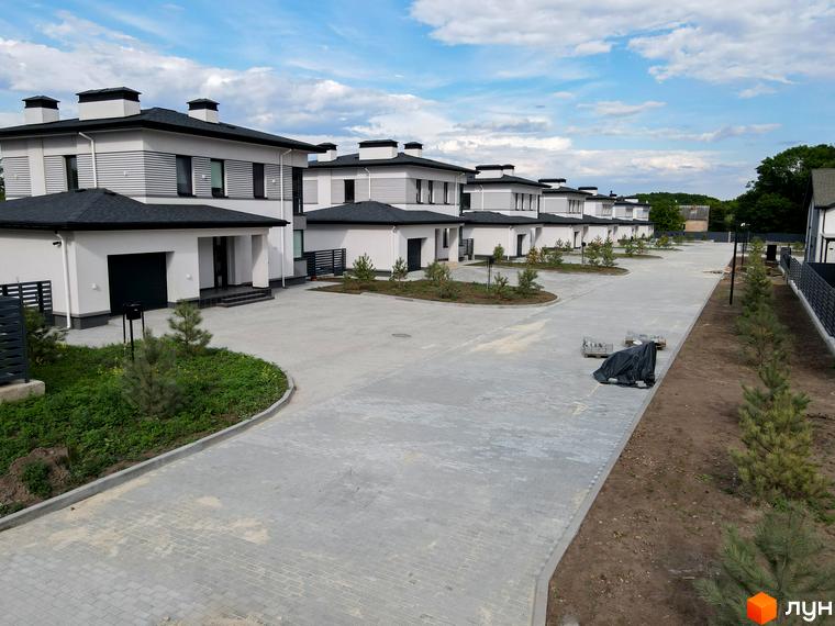 Котеджне містечко Ecopark: ряд двоповерхових будинків з білими фасадами та темними дахами. Брукована дорога та молоді дерева на території.