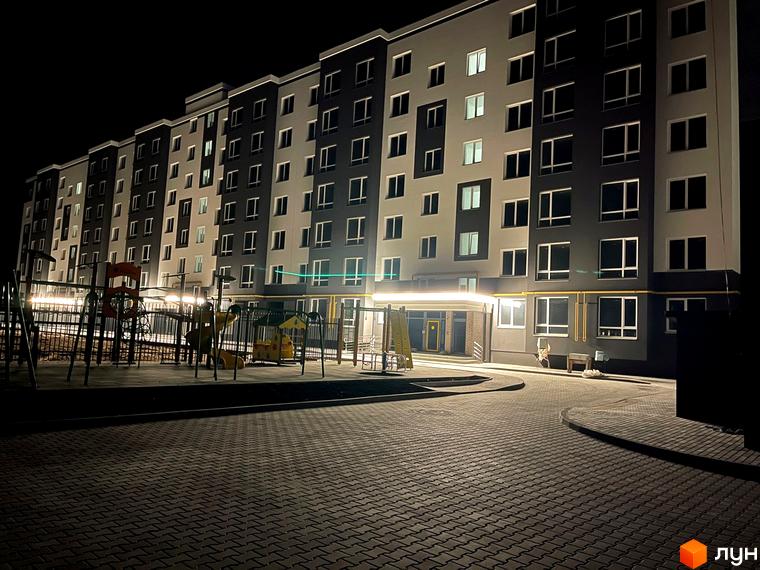 Новобудова ЖК Петрівські Липки з фасадом у сірих та білих тонах, освітленим вхідним групами, та дитячим майданчиком поруч.