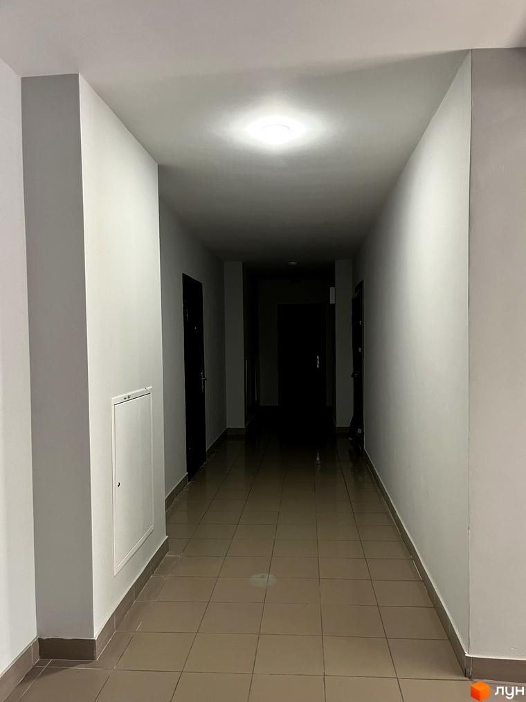 Коридор з білими стінами та світлою плиткою на підлозі. У кінці коридору видно темні двері.