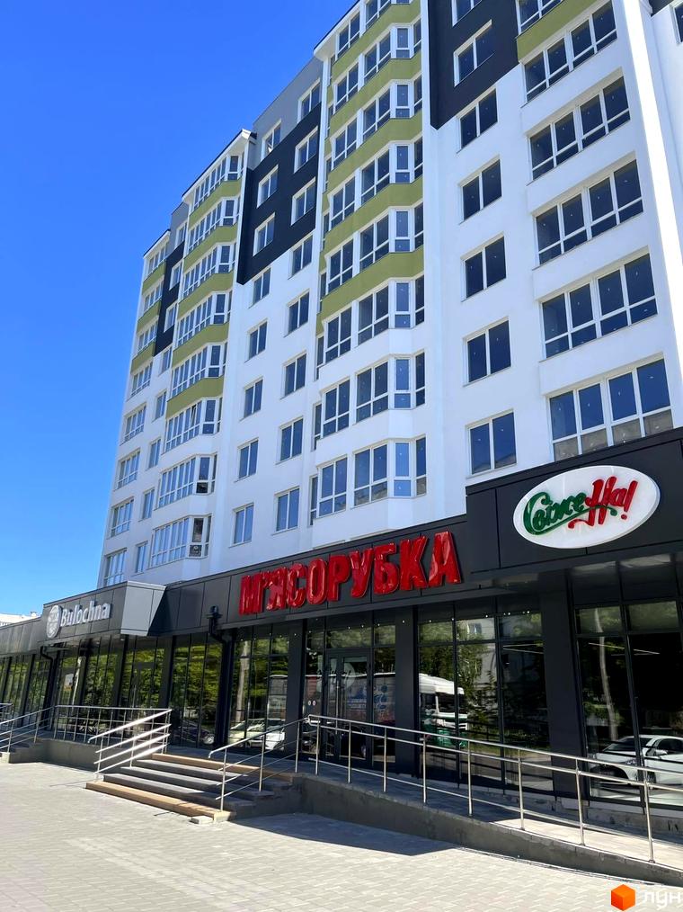 Висотна будівля з великою кількістю вікон та магазинами на першому поверсі. Біля будинку розташована доглянута прибудинкова територія з тротуаром та сходами.