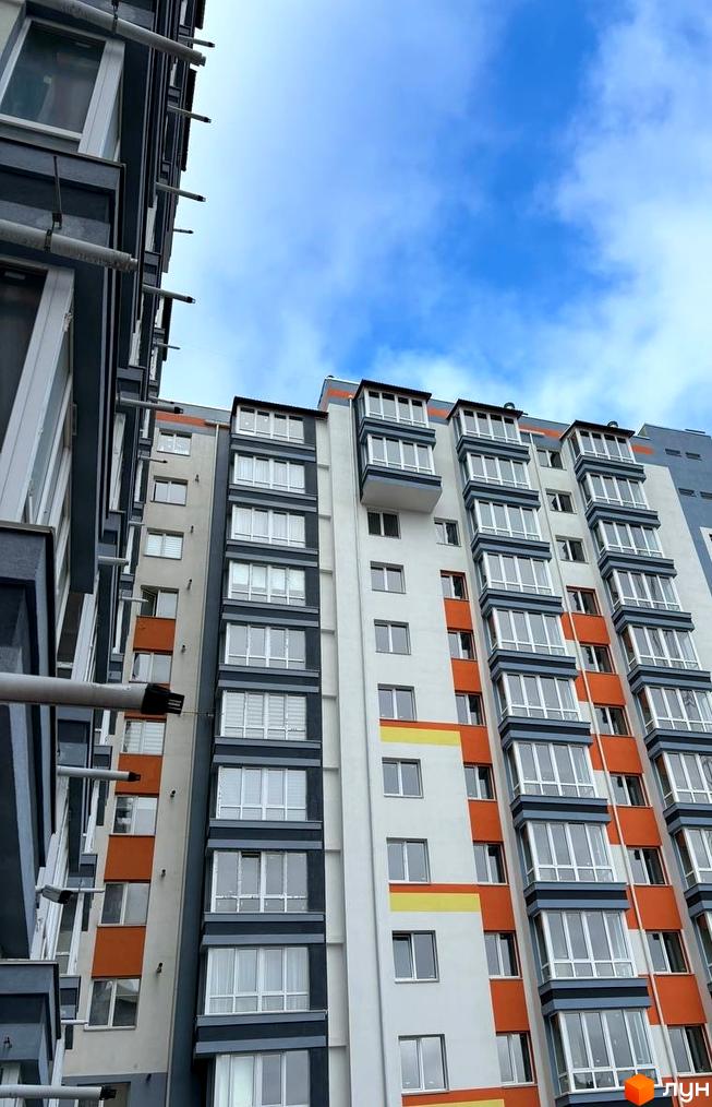 Фасад багатоповерхового будинку МЖК Сім'я з білими, сірими та помаранчевими секціями, заскленими балконами під блакитним небом.
