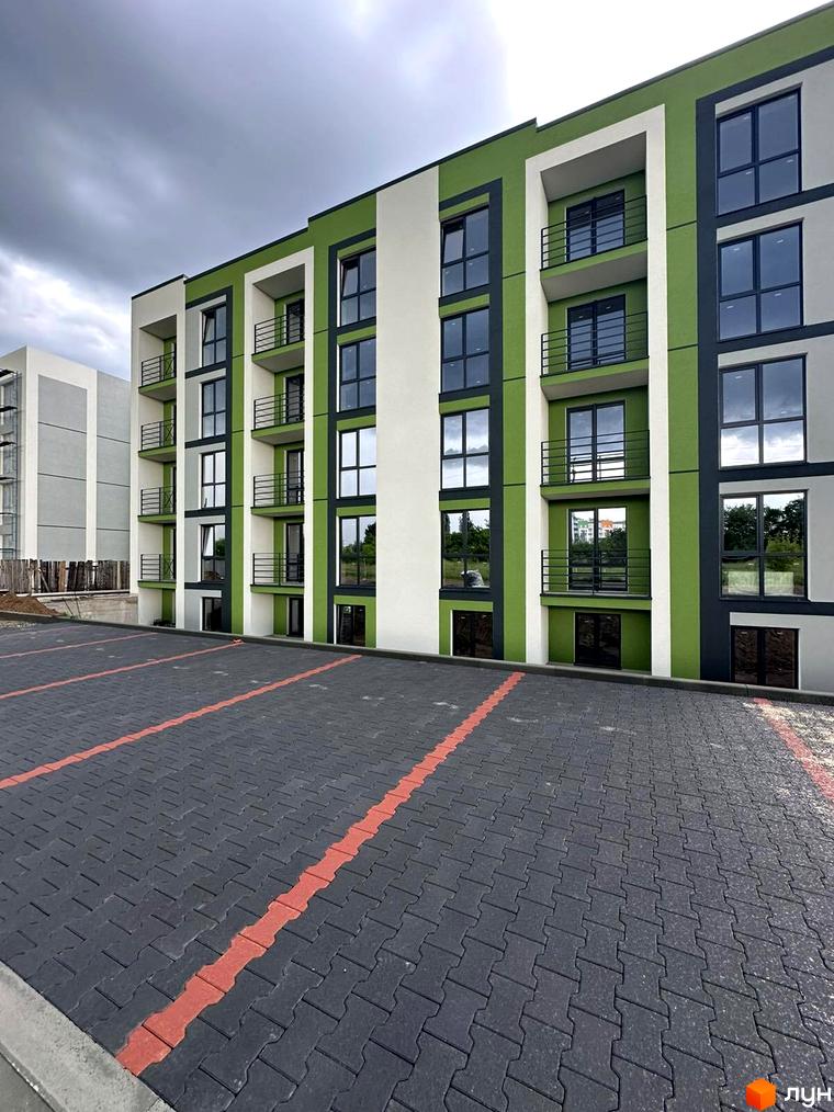 Житловий комплекс Brickwood 2 з яскравим фасадом у зелених та білих тонах, з балконами та великими вікнами. Поруч розташований паркінг з червоними розмітками.