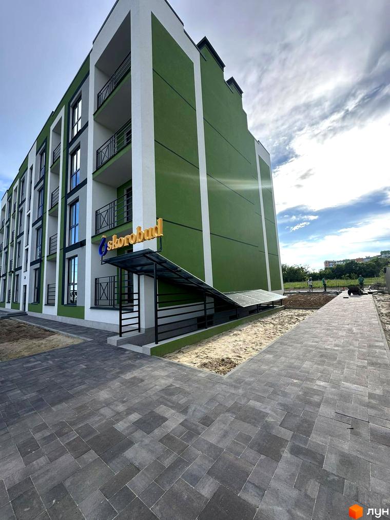 Зовнішній вигляд новобудови ЖК Brickwood 2 з зеленими та білими фасадами, балконами та бруківкою на прибудинковій території.